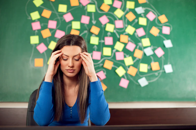 Teacher sitting at desk with her eyes closed, with map of different coloured posit notes on chalkboard behind her.
