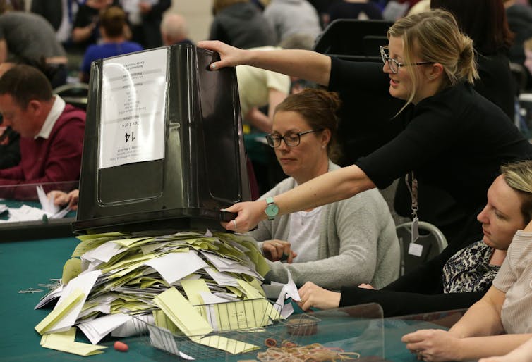 Woman empties container of ballot papers on table in front of other women.