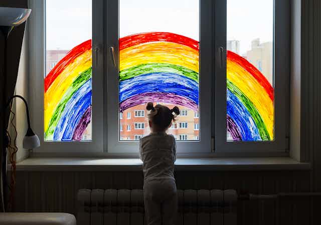 Una niña asomada a una ventana en cuyos cristales hay pintado un arcoriris.