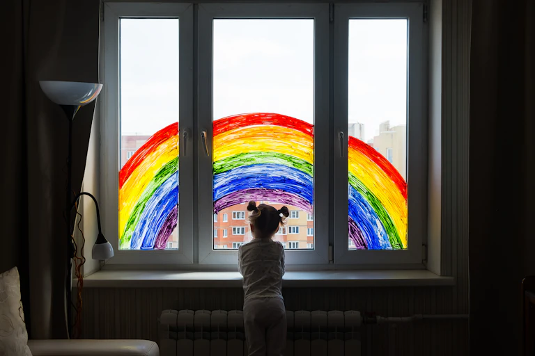 Una niña asomada a una ventana en cuyos cristales hay pintado un arcoriris.