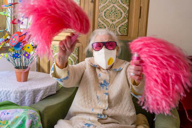 An elderly woman wears a mask while waving pink pompoms.