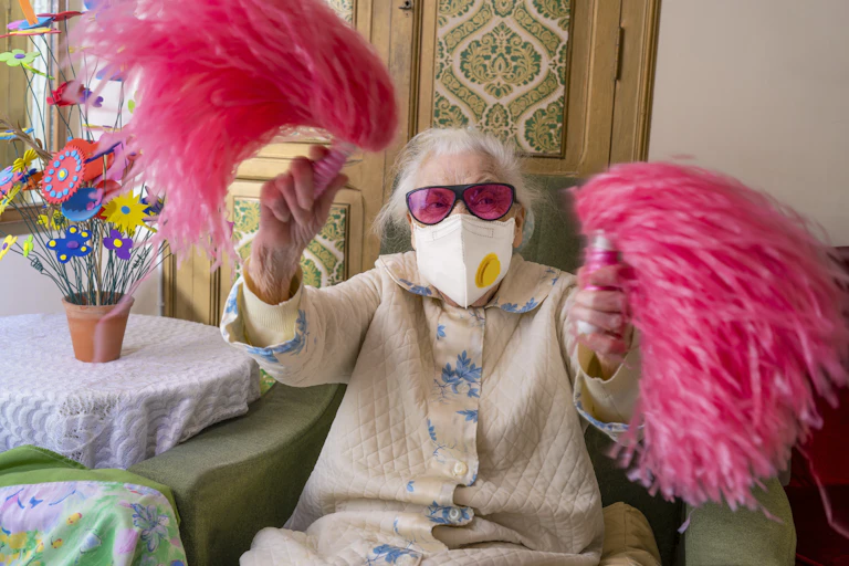 An elderly woman wears a mask while waving pink pompoms.