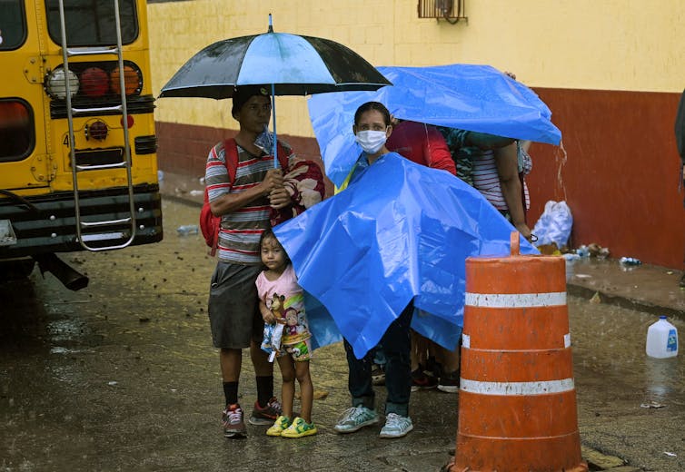 A family stands in the rain under umbrellas and tarps, behind a school bus