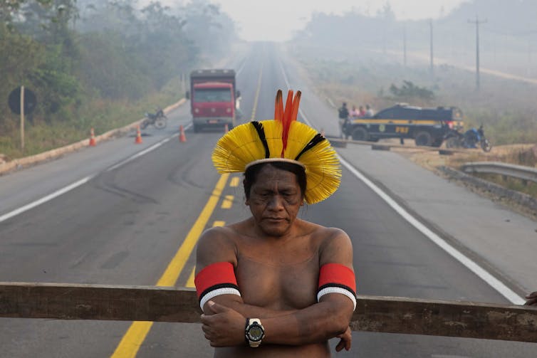 An indigenous man stands in front of a roadblock.