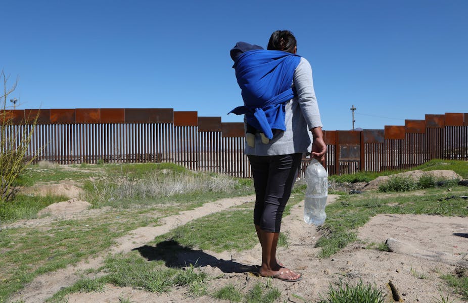 A migrant carrying a toddler on her back stands in front of the border wall that divides the US and Mexico 