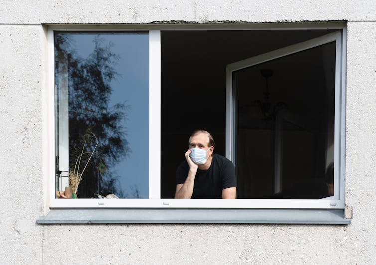 A man wearing a mask looks out the open window of his home.