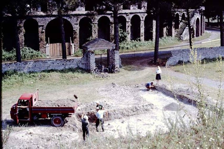 A red ute and five people working in a shallow ditch outside an amphitheatre.