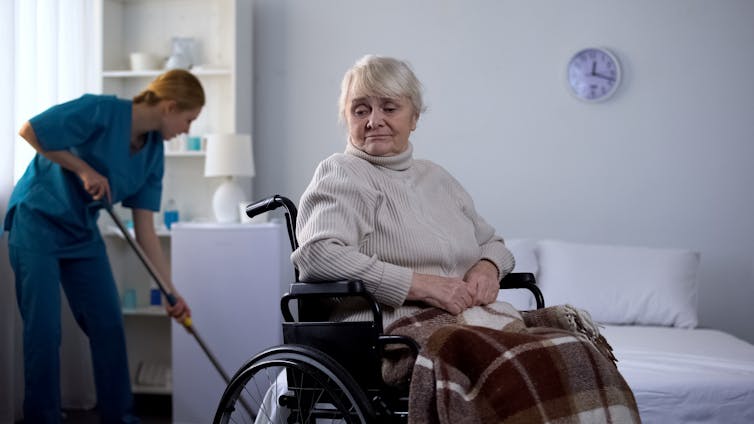 An elderly woman sits in a wheelchair, while a cleaner cleans the floor of her room.