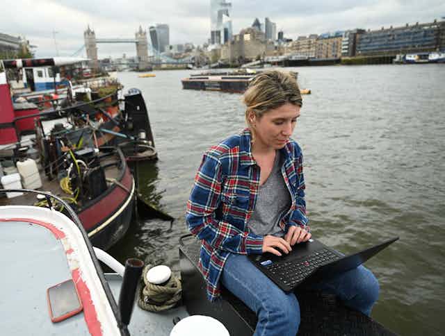 A woman sits with a laptop on her house barge on the Thames river.