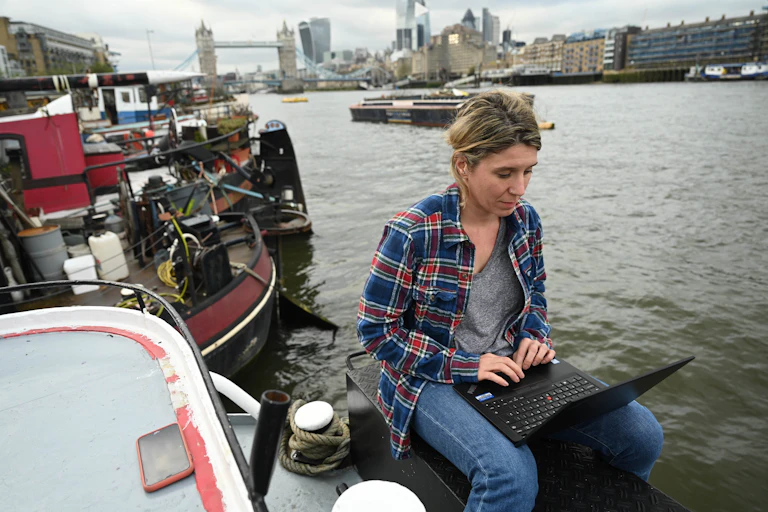 A woman sits with a laptop on her house barge on the Thames river.
