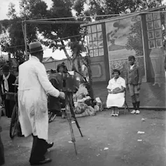 A man takes a photograph of a woman and her son on the streets of Cape Town. They stand against a painted backdrop of a balcony overlooking the sea, with a volcano erupting in the distance.