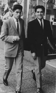 Two men walk along a pavement, both dapper in suits, the one looking at the camera questioningly and the other smiling at it broadly.