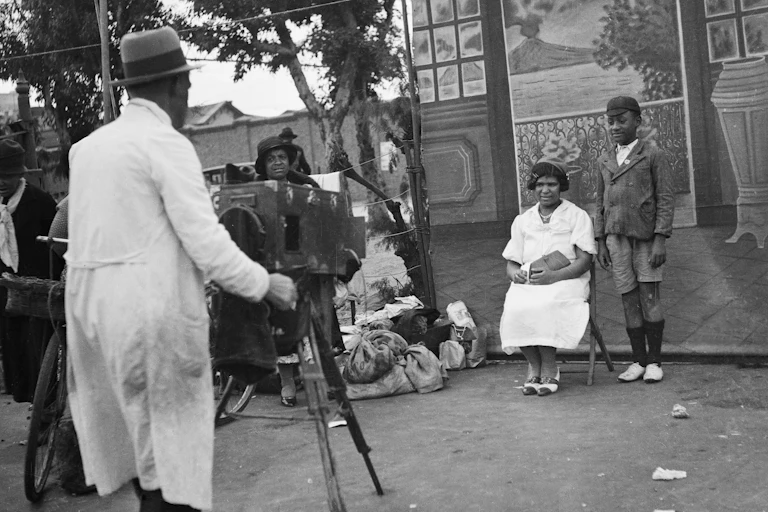 A man in a hat and coat photographs a woman and boy against a backdrop of a smart home and a volcano erupting out to see. Rubbish and litter can be seen on the street, where a woman looks on.