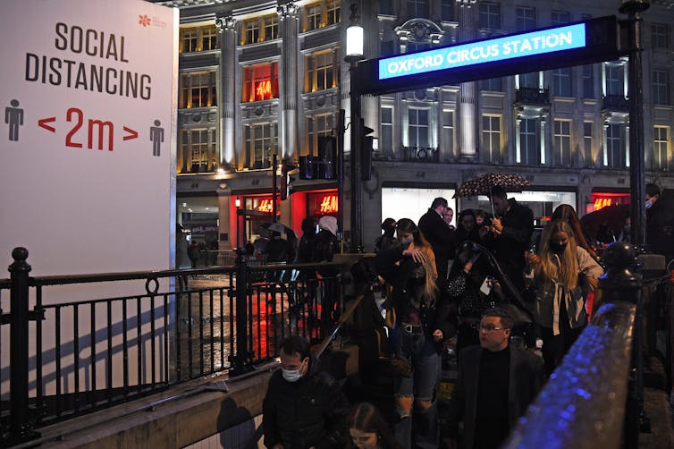 A crowd of people enters Oxford Circus underground station next to a sign warning them to maintain a social distance of 2 metres.