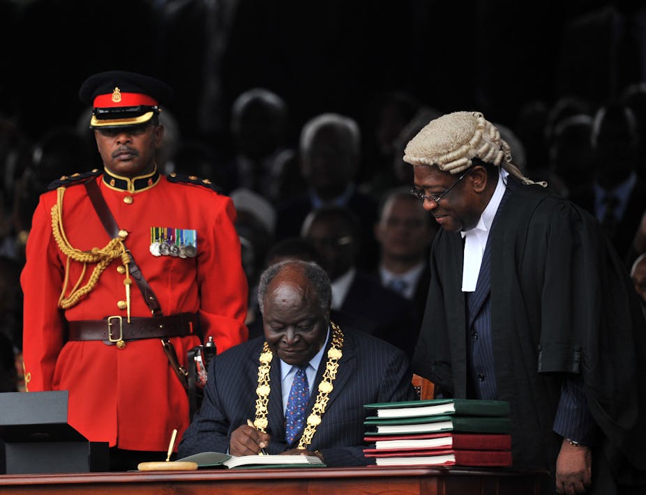 Former President Mwai Kibaki, seated and wearing a gold presidential chain, signs the constitution, watched by the former Attorney General and a soldier dressed in red uniform.
