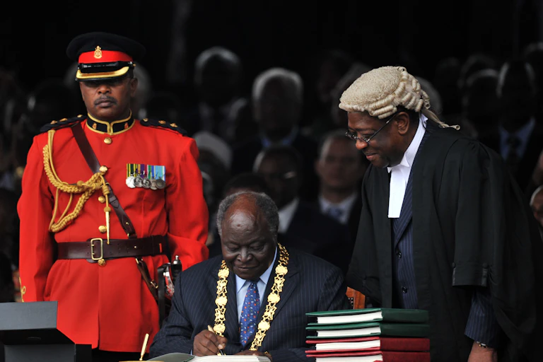Former President Mwai Kibaki, seated and wearing a gold presidential chain, signs the constitution, watched by the former Attorney General and a soldier dressed in red uniform.