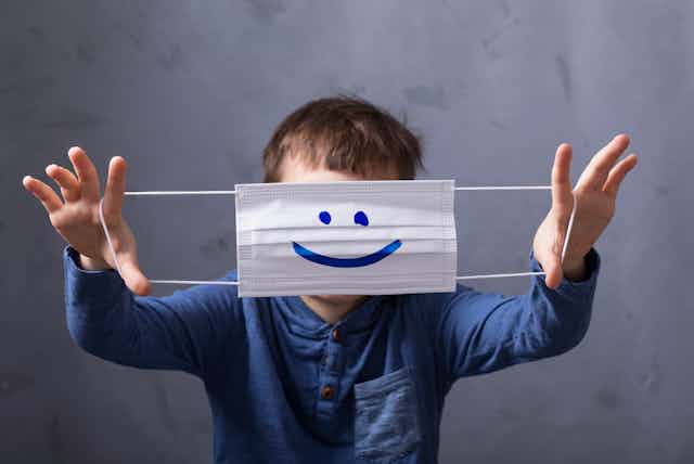 Un niño se tapa la cara mostrando una mascarilla con una sonrisa dibujada.