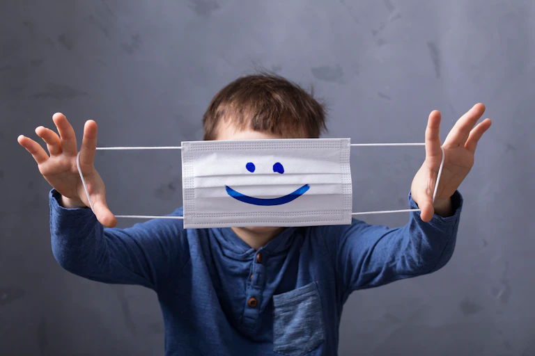 Un niño se tapa la cara mostrando una mascarilla con una sonrisa dibujada.