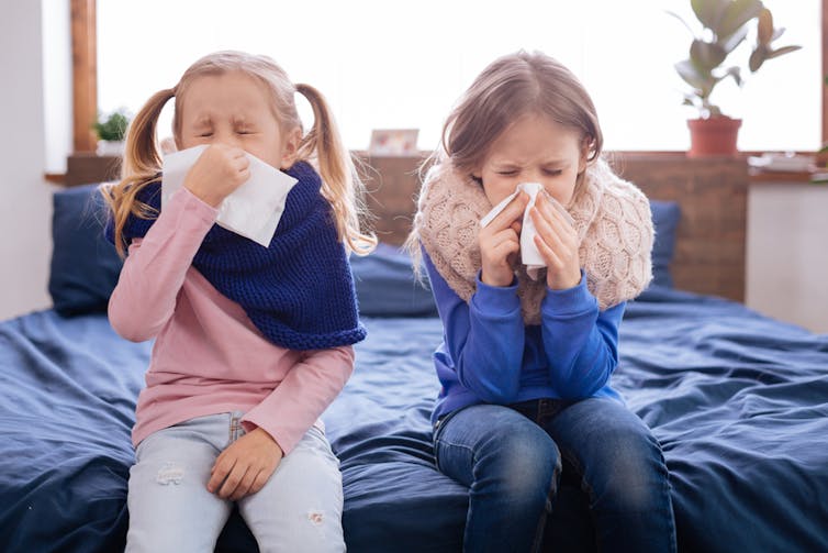 Two girls sit on the edge of a bed blowing their nose.