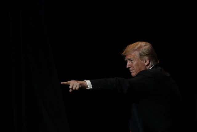 President Donald Trump, surrounded by darkness, is seen pointing to the left of the image.