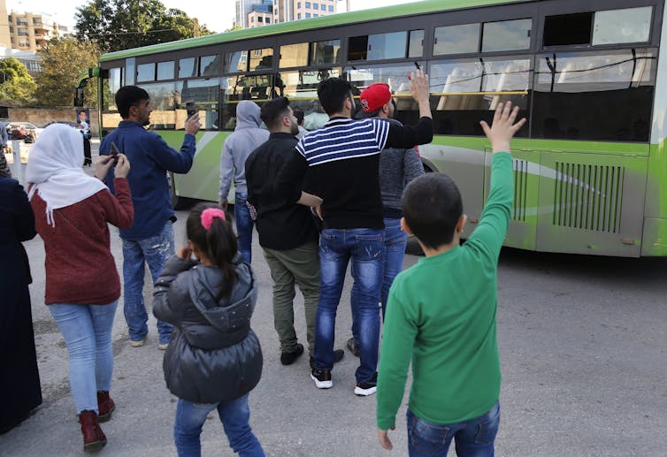 People stand near a green bus waving to its occupants.