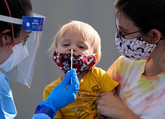 A health-care worker in PPE is about to insert a long swab into a little blond boy's nose as his mother holds him. The boy and his mother are wearing cloth masks.