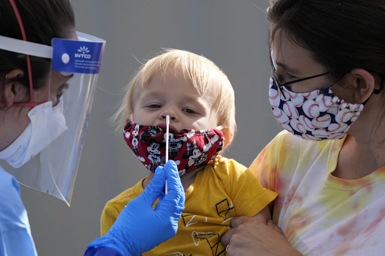 A health-care worker in PPE is about to insert a long swab into a little blond boy's nose as his mother holds him. The boy and his mother are wearing cloth masks.