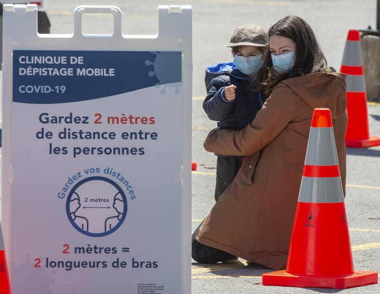 A masked woman hugs her masked child between orange pylons behind a sign for a mobile testing facility that instructs people to keep two metres apart.