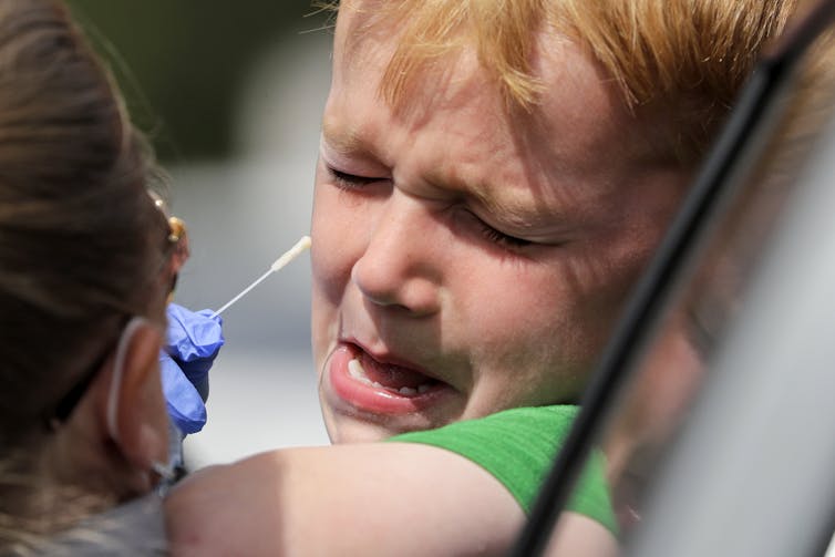 A child sticks his head out a car window and winces as a health-care worker is about to insert a swab in his nose.