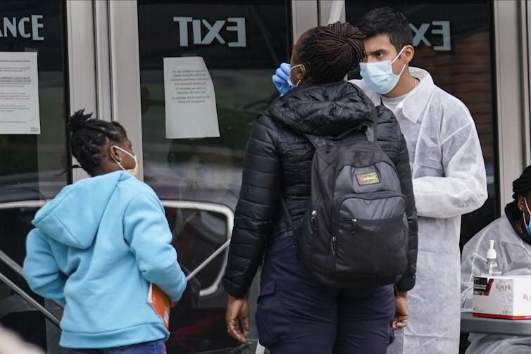 A white-coated health-care worker swabs a woman's nose while a girl watches.