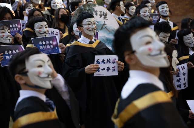 Crowd of students in graduation robes wearing Guy Fawkes masks, holding signs.