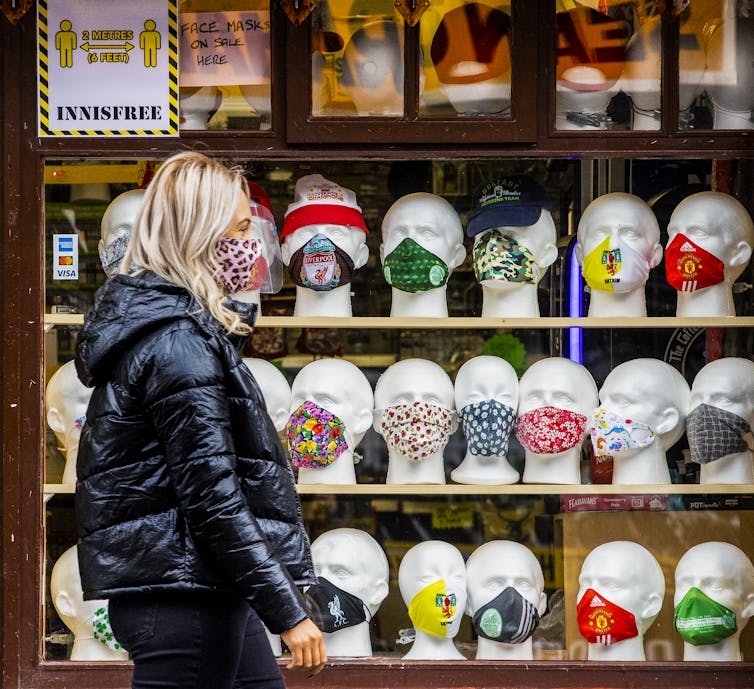 A woman wearing a mask walks past a shop window containing many mannequin heads wearing masks.