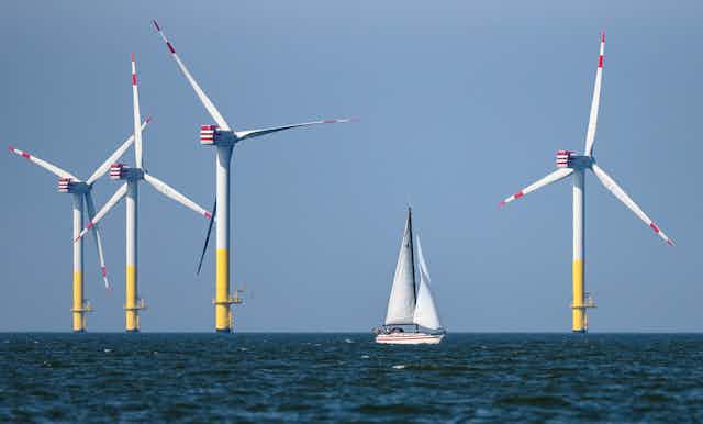 Four offshore wind turbines stand amid the North Sea with a white sail boat in the foreground.