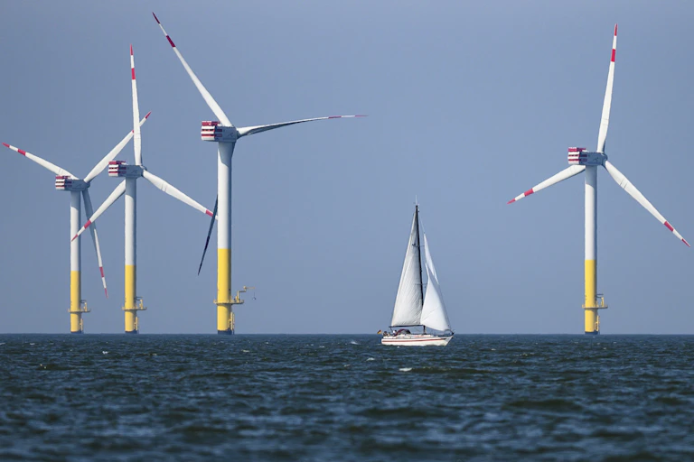 Four offshore wind turbines stand amid the North Sea with a white sail boat in the foreground.