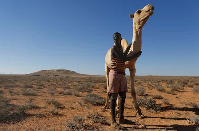 Young man poses with camel in desert scrub landscape.