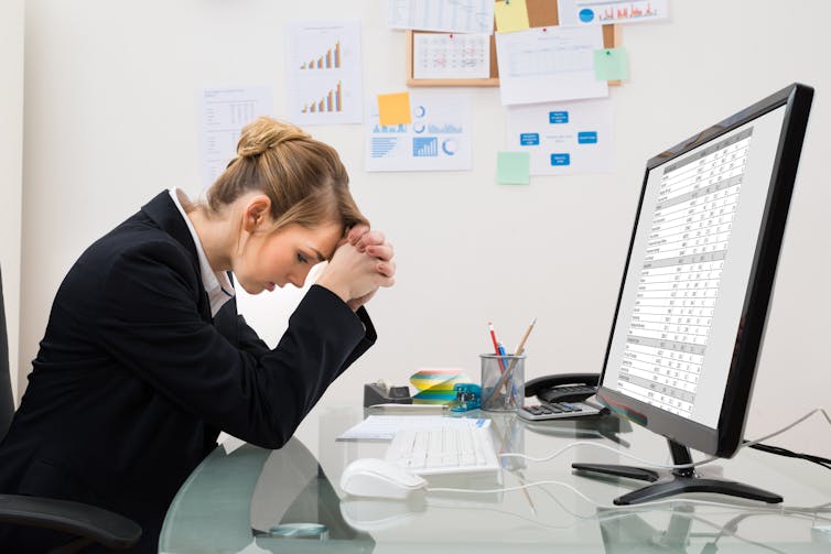 Woman at computer with spreadsheet on screen and head in hands.