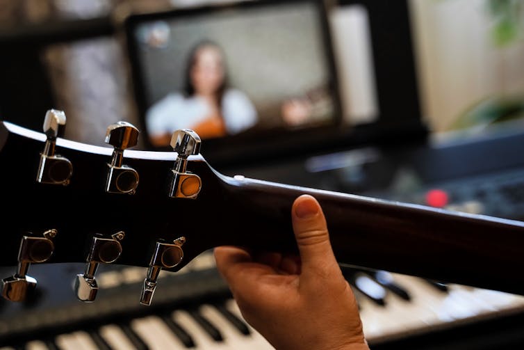 Arm of person playing guitar, with another person playing on screen in background.