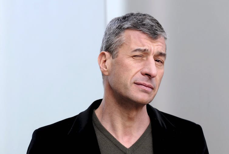 Maurizio Cattelan winking in black blazer.