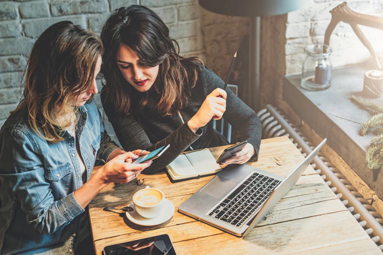 Two young women seated at a table in front of a laptop and looking at phones