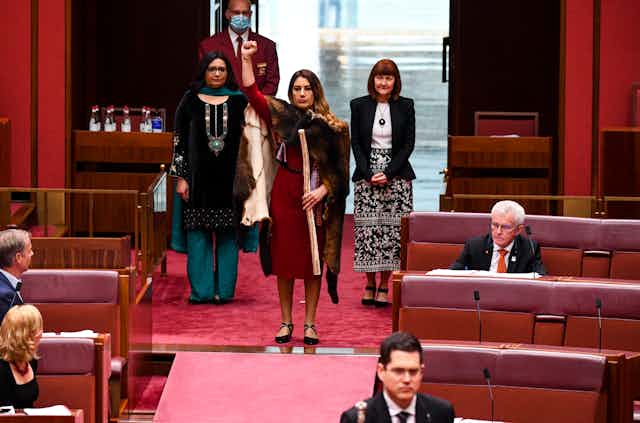 Woman in Senate chamber of Parliament with fur cloak, fist raised, holding message stick. 
