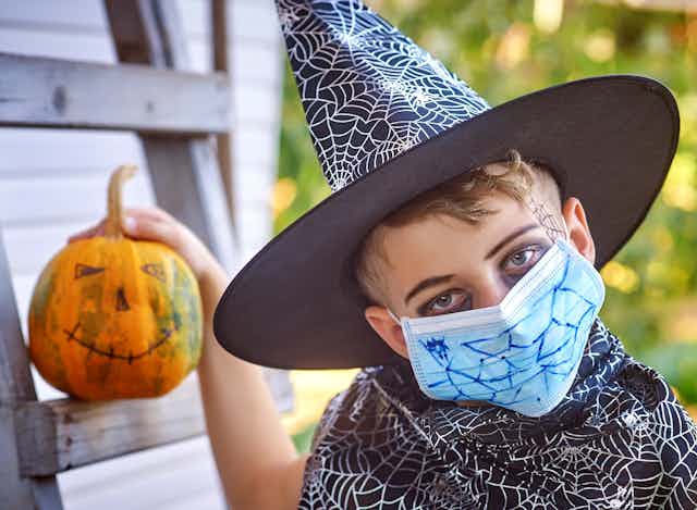 A young boy wearing a black witches hat with a surgical mask on his face holding a painted Halloween pumpkin