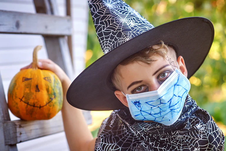 A young boy wearing a black witches hat with a surgical mask on his face holding a painted Halloween pumpkin