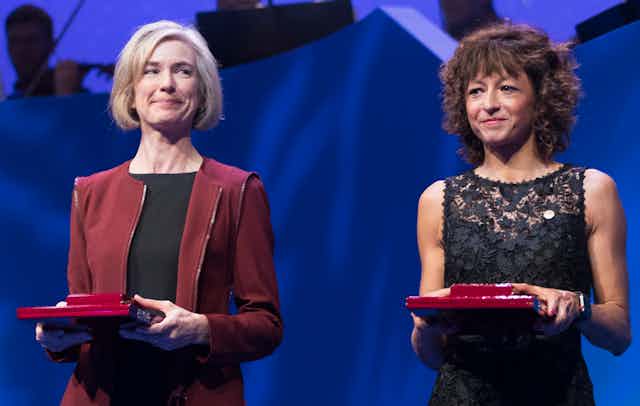 Jennifer Doudna and Emmanuelle Charpentier in smart dress holding red prize boxes.