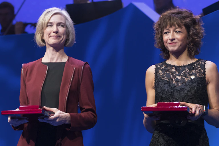 Jennifer Doudna and Emmanuelle Charpentier in smart dress holding red prize boxes.