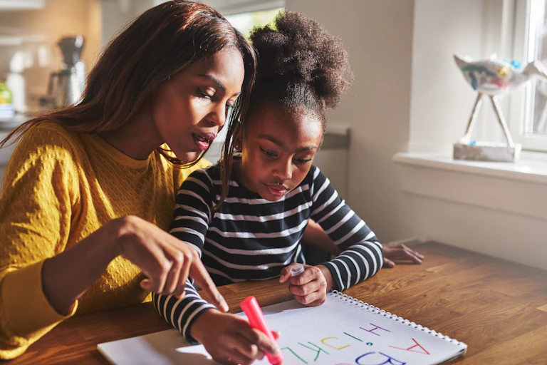 A well groomed woman in a gold sweater supervises her daughter as she does homework using a red marker and drawing paper.