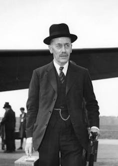 Man in three-piece suit and hat carrying briefcase and umbrella with aircraft in the background.