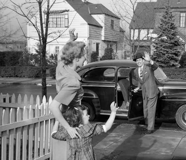 Mother and child waving goodbye to father who is heading to work in 1950's suburb.