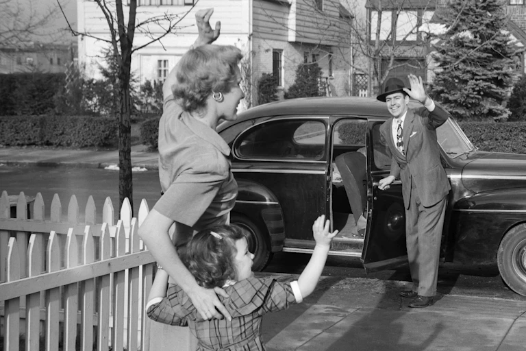 Mother and child waving goodbye to father who is heading to work in 1950's suburb.