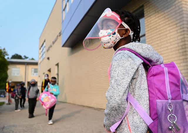 A young girl wears a mask and face shield as she waits in line for her kindergarten class to enter her school.