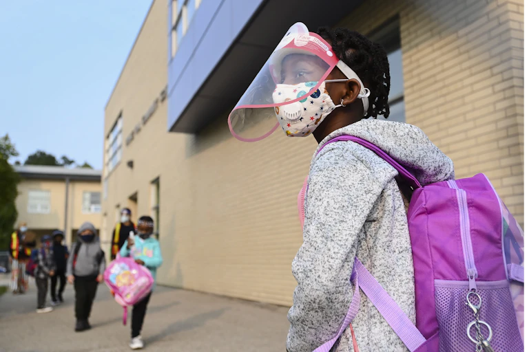 A young girl wears a mask and face shield as she waits in line for her kindergarten class to enter her school.